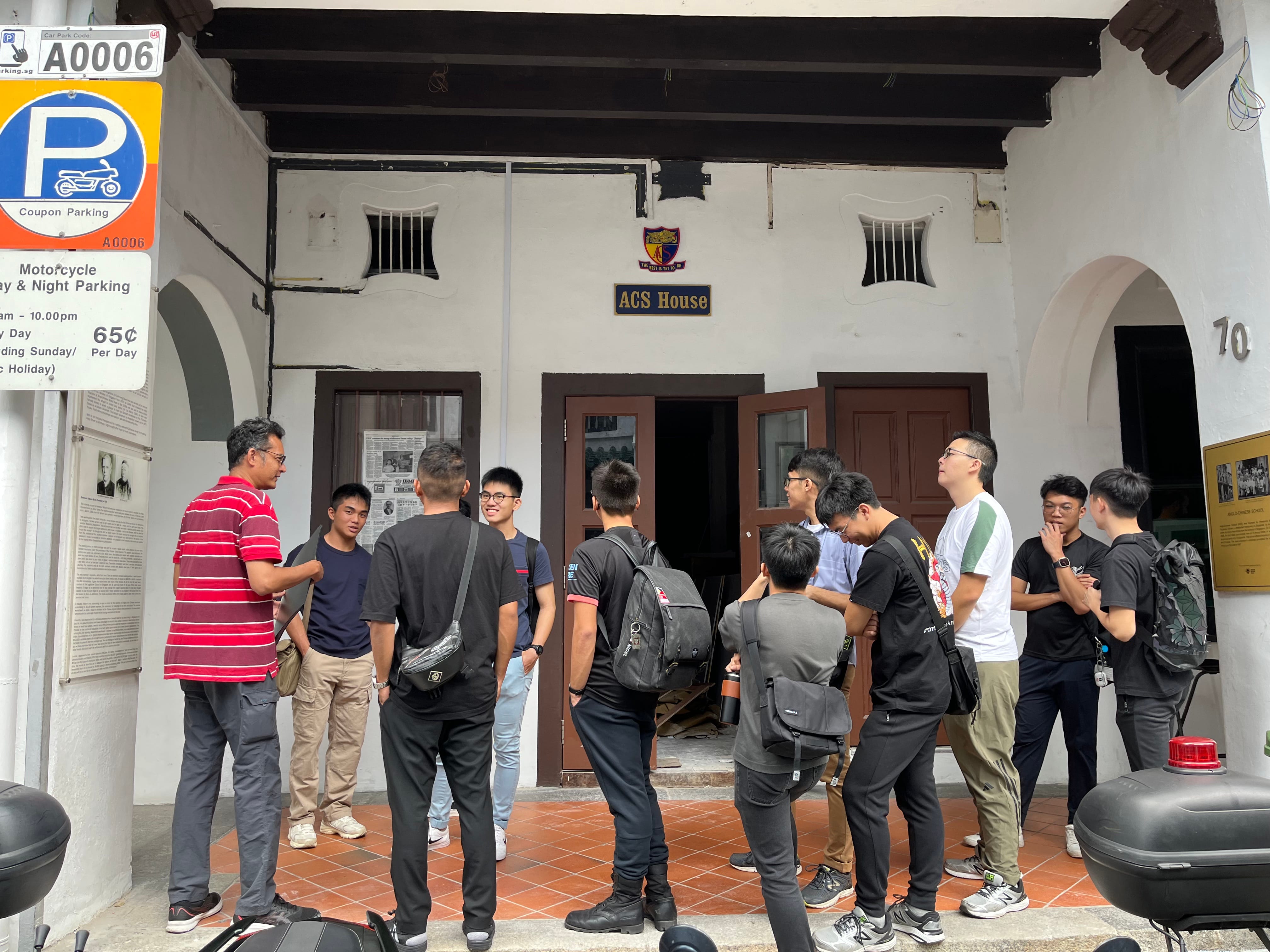 A group of young people stand outside a white colonial building labeled ACS House, with a parking sign nearby.